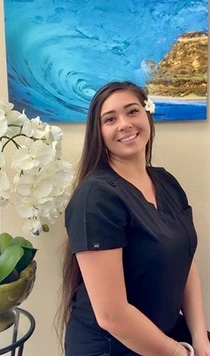 Dental assistant in black scrubs with flower in hair, smiling by orchid and ocean art at dental office.